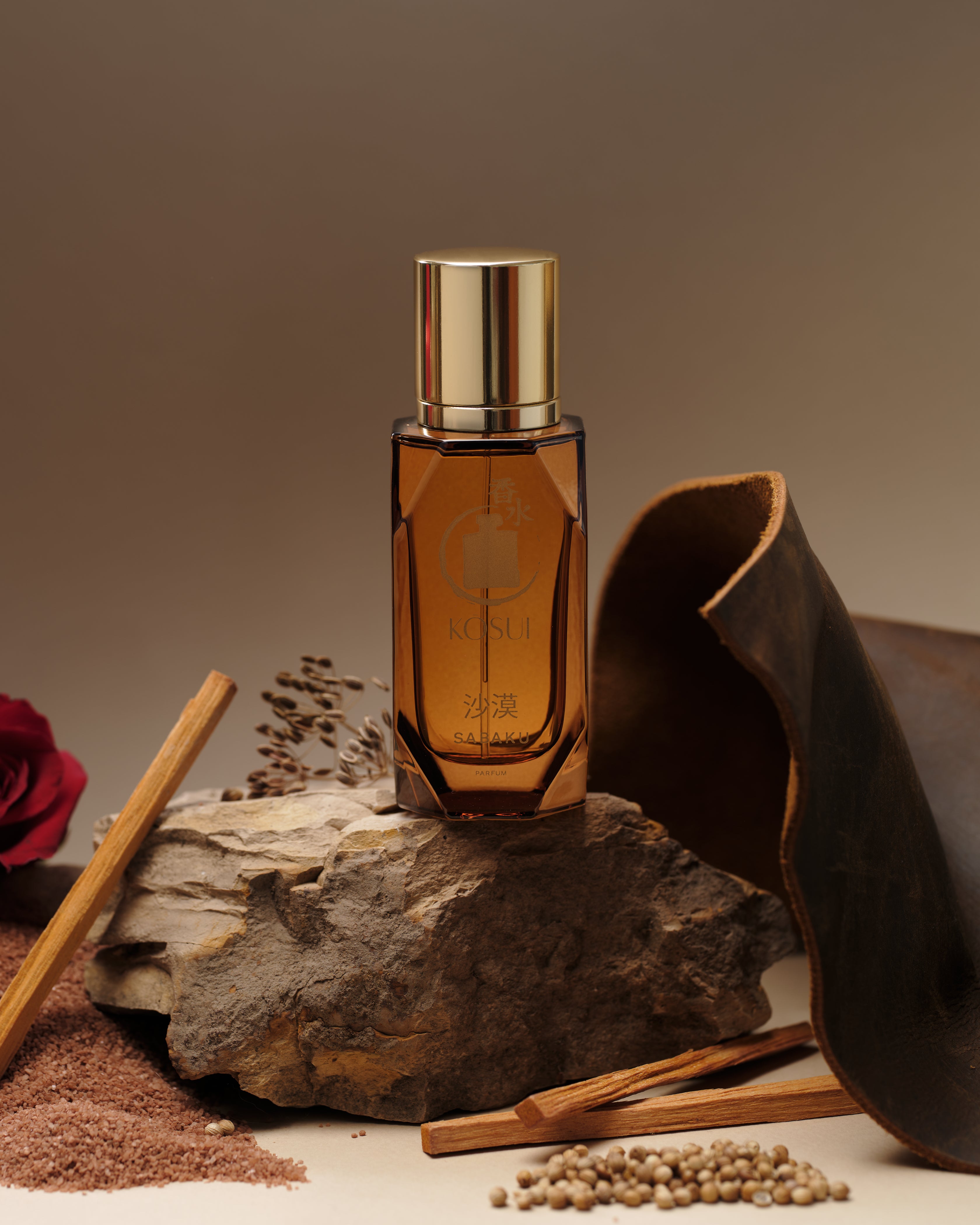 Brown perfume bottle with gold cap on a stone surface with cinnamon sticks and a rose.